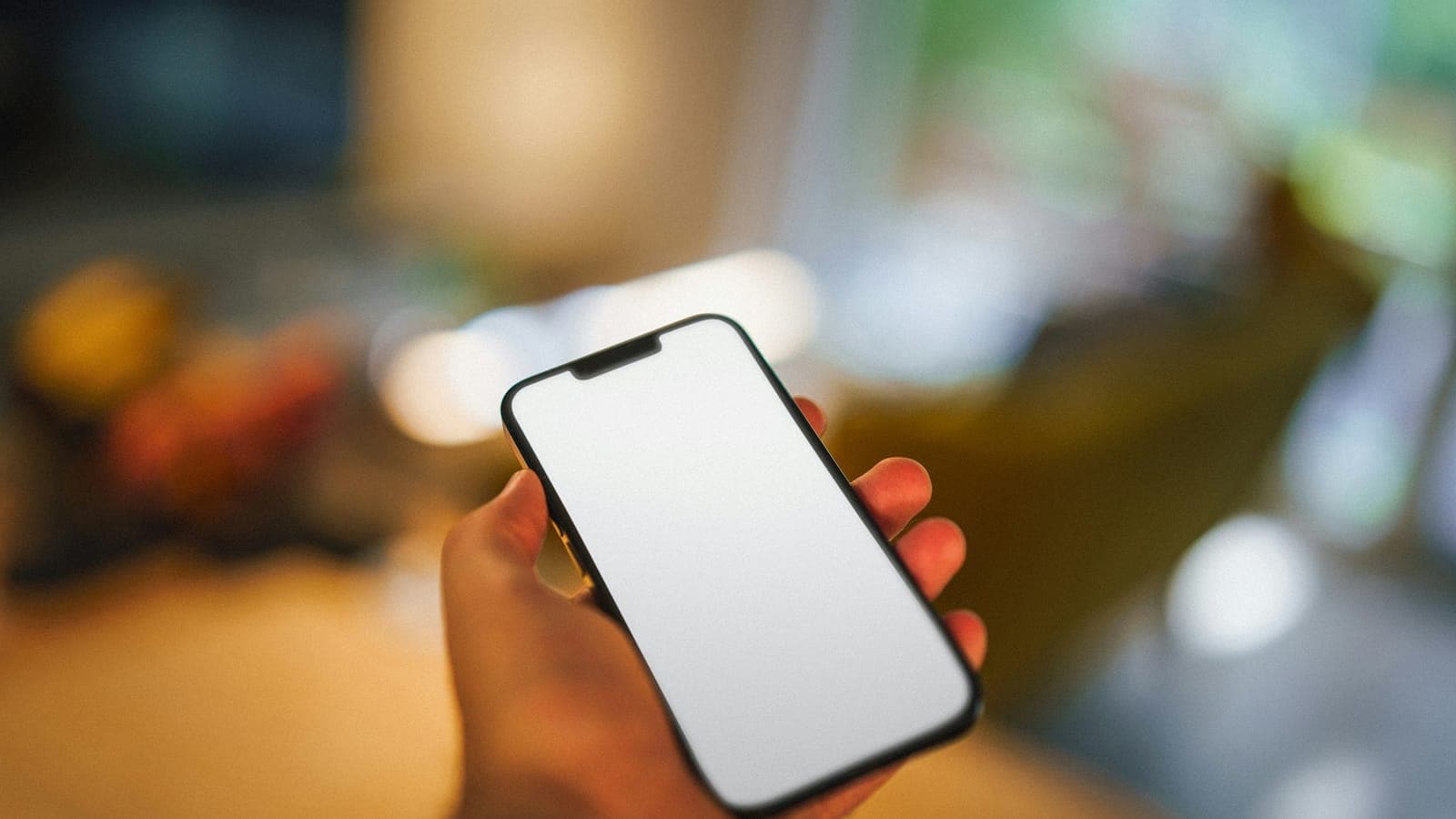 Mano sosteniendo un smartphone con la pantalla en blanco sobre un fondo desenfocado de tonos cálidos